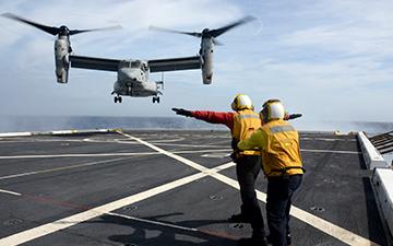 Two people watching a MV-22 take off  from an aircraft carrier